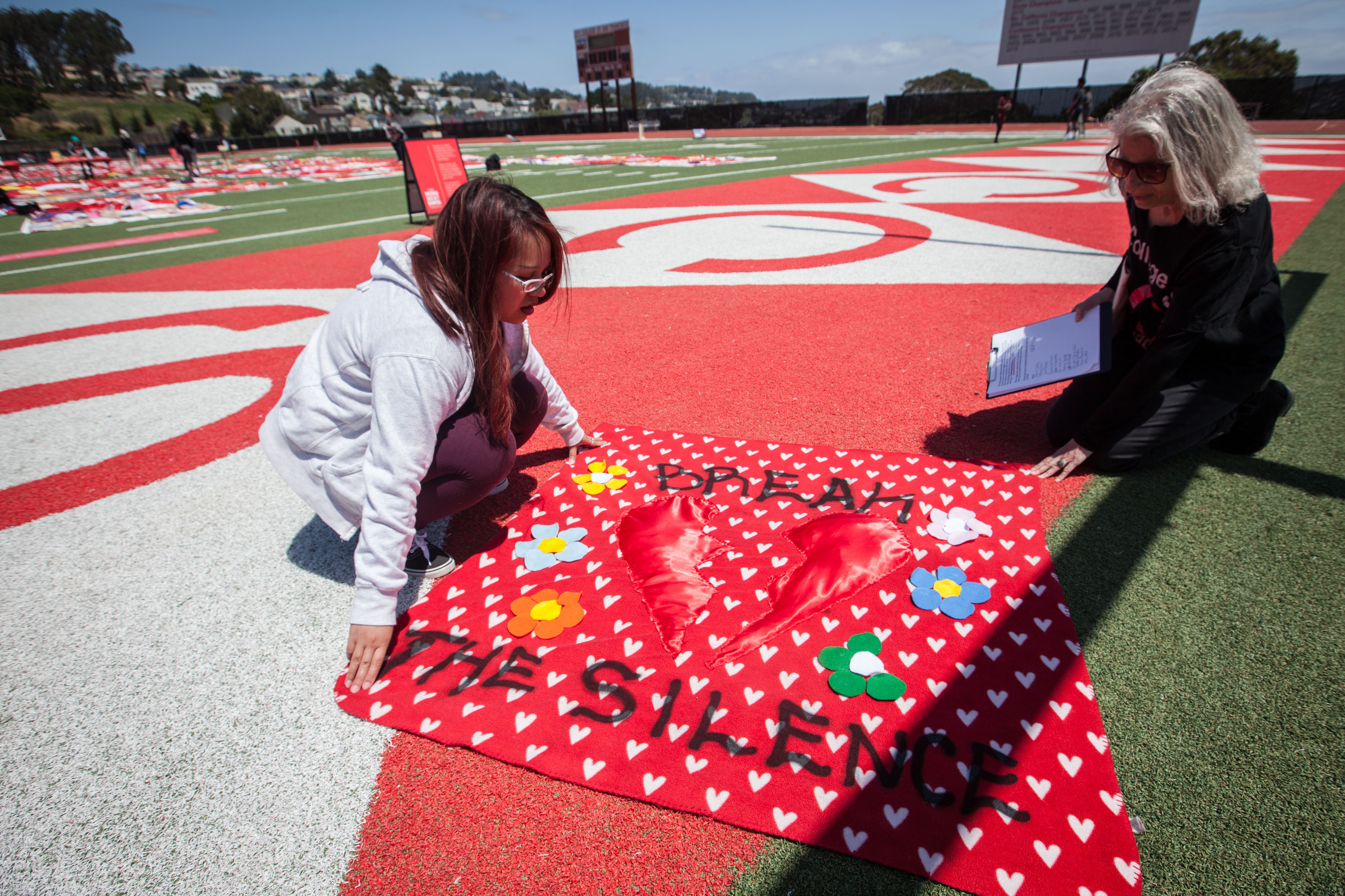 (L-R) An instructor of Ending Sexual Violence: Peer Education Shella Cervantes and Interim Project SURVIVE Coordinator Leslie Simon display a red square quilt made by a CCSF student Valentina Vargas during an installation of The Monument Quilt on the football field of George M. Rush Stadium at City College of San Francisco Ocean Campus on Saturday, May 6, 2017. Vargas made the quilt out of her old blanket that she had since she was 16 years old and figured that the blanket had seen all her past unhealthy relationship; so making it into a quilt is the way for her to recycle old energy through healing and art. (Photo by Ekevara Kitpowsong) 